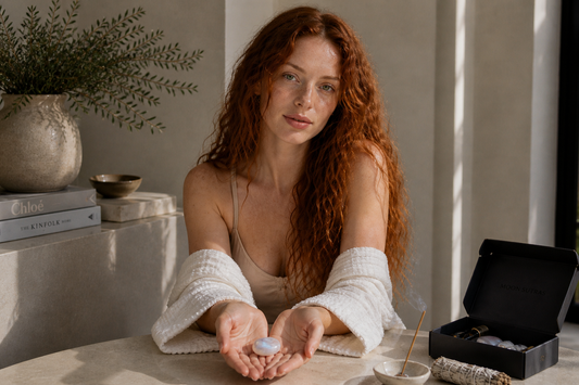 Moonstone palm stone held in both hands by red-haired woman with freckles at sunlit table, Moon Sutras ritual box and incense nearby