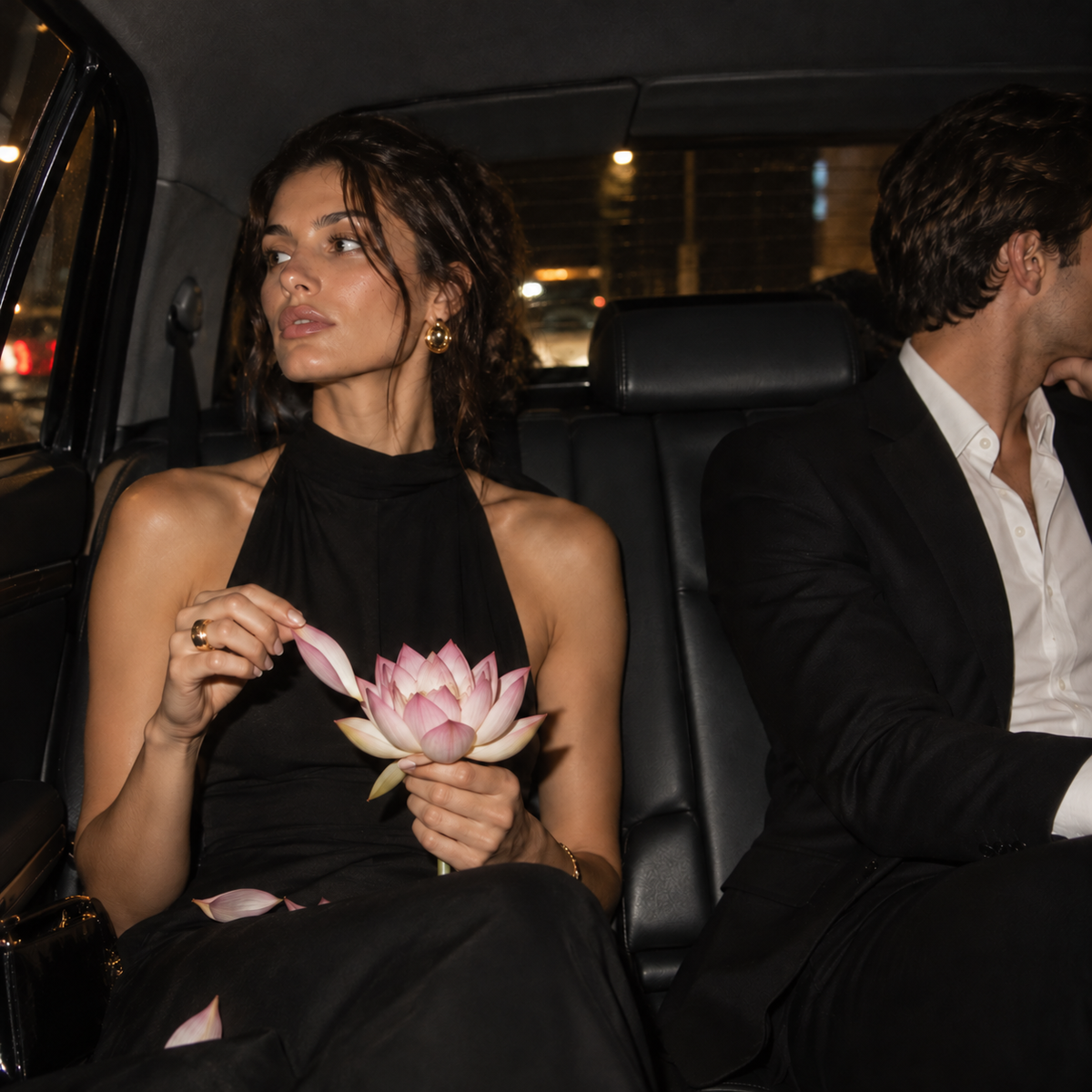 Woman in a black dress removing petals from a pink lotus in the back of a car at night, while a man in a black suit looks away.