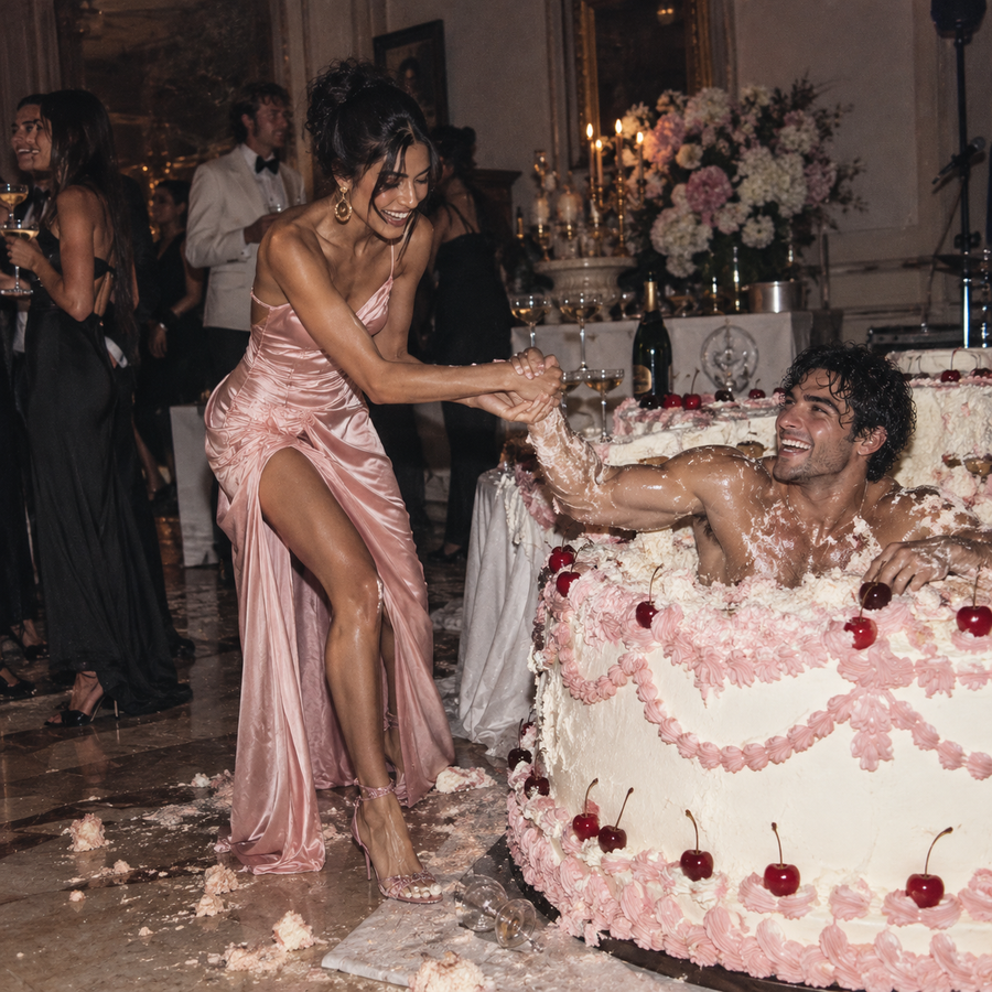 Woman in a pink satin evening dress laughing as she helps a bare-chested man covered in icing climb out of an enormous birthday cake at a lavish party.