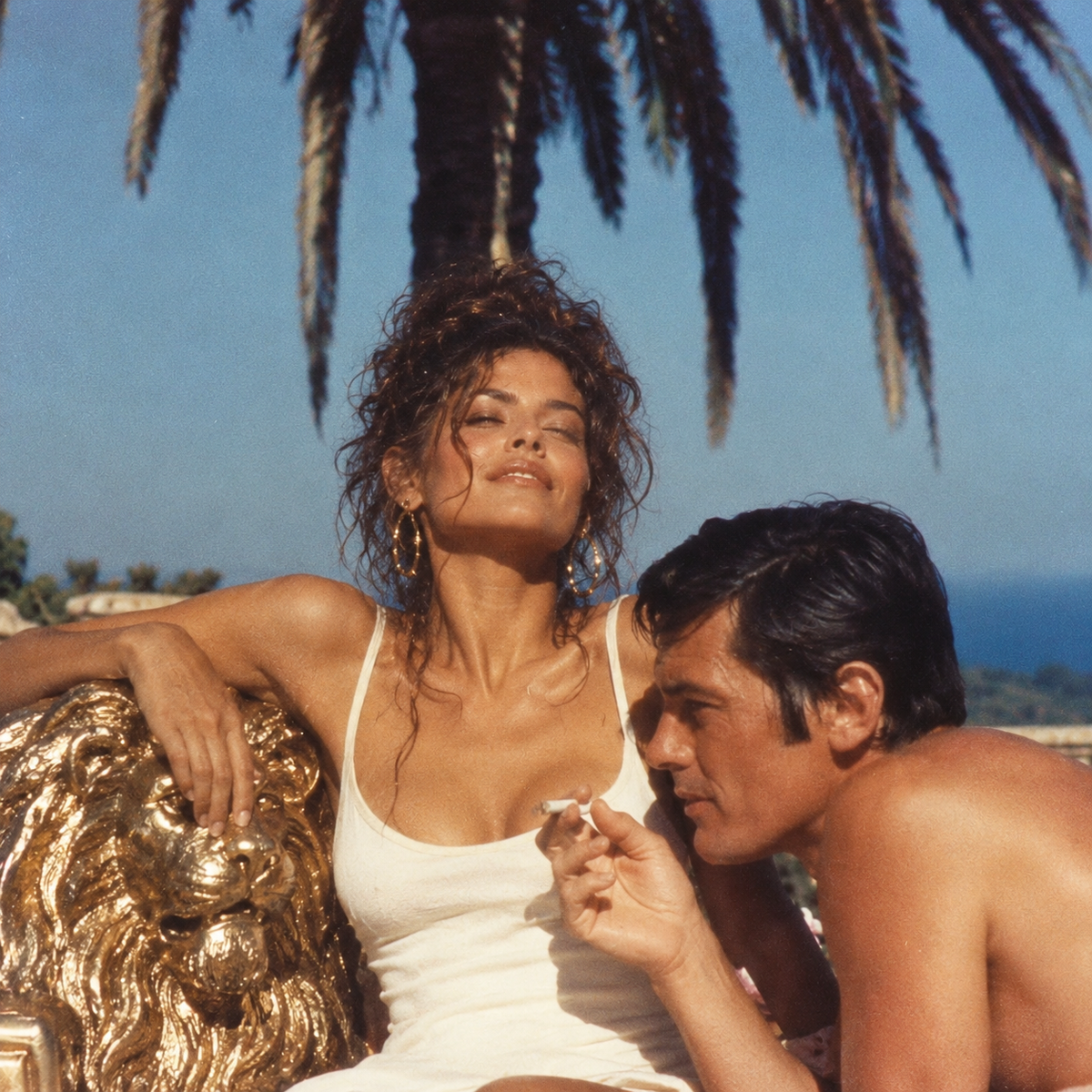 Glamorous brunette with a tousled updo and gold hoop earrings reclines beside a golden lion throne, wearing a white swimsuit, while a dark-haired man leans beside her beneath palm trees in a grainy 1990s Mediterranean fashion photograph.
