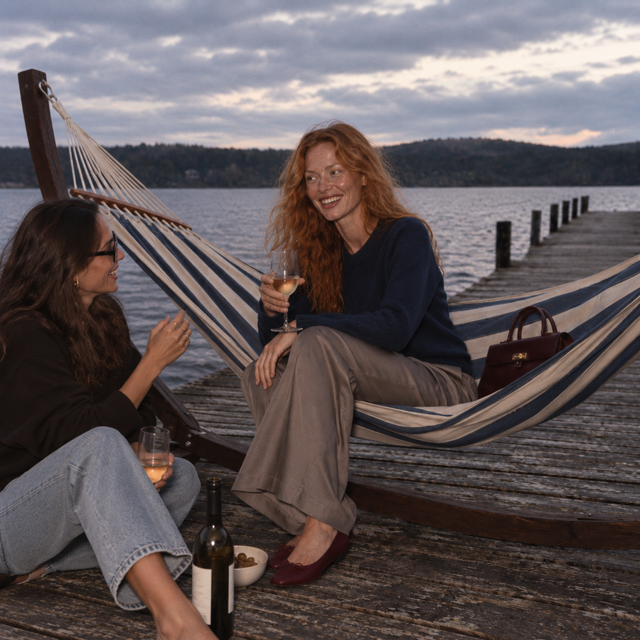 Freckled red-haired woman in a navy jumper and taupe trousers laughing with a friend in sunglasses beside a striped hammock on a lakeside jetty.