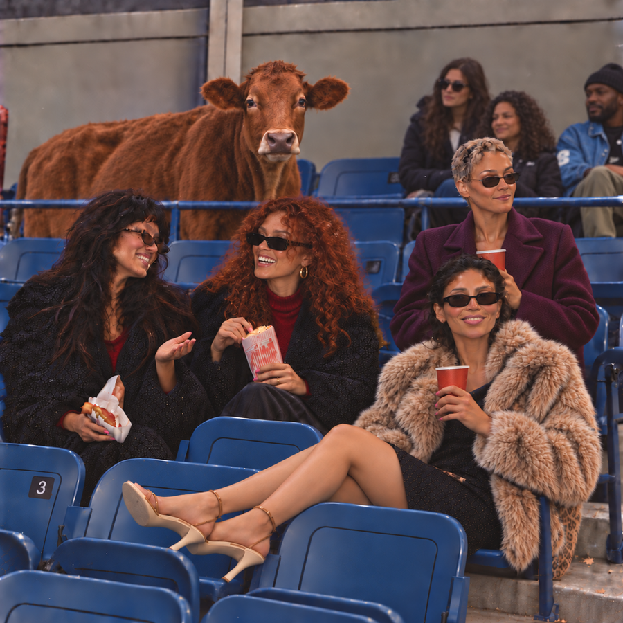 Pushya Nakshatra editorial photograph of four fashionable women in sunglasses and winter coats enjoying popcorn and drinks in stadium seats, with a brown cow standing behind them, reflecting Pushya's symbolism of nourishment and care in Vedic astrology.