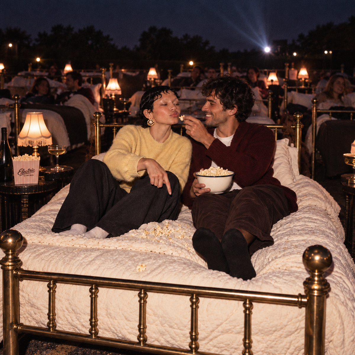 Couple sharing popcorn on a brass bed at an outdoor cinema, surrounded by other filmgoers reclining on beds beneath warm vintage lamps.